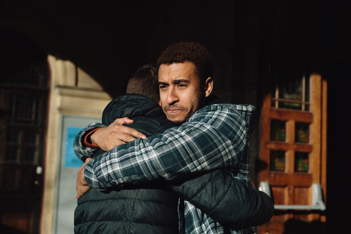 A man looking sad while hugging another man outside a doorway.