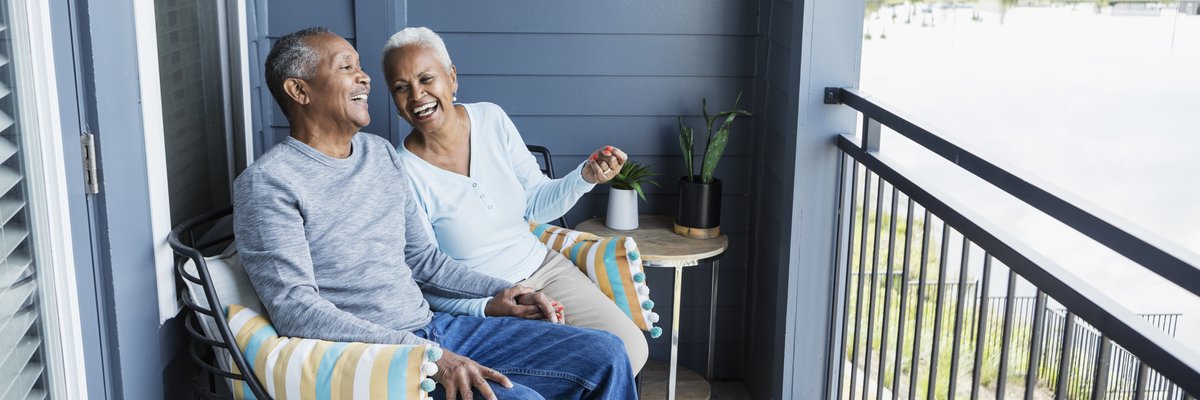 Two mature adults laughing together while sitting on the back deck of their home overlooking a small lake.
