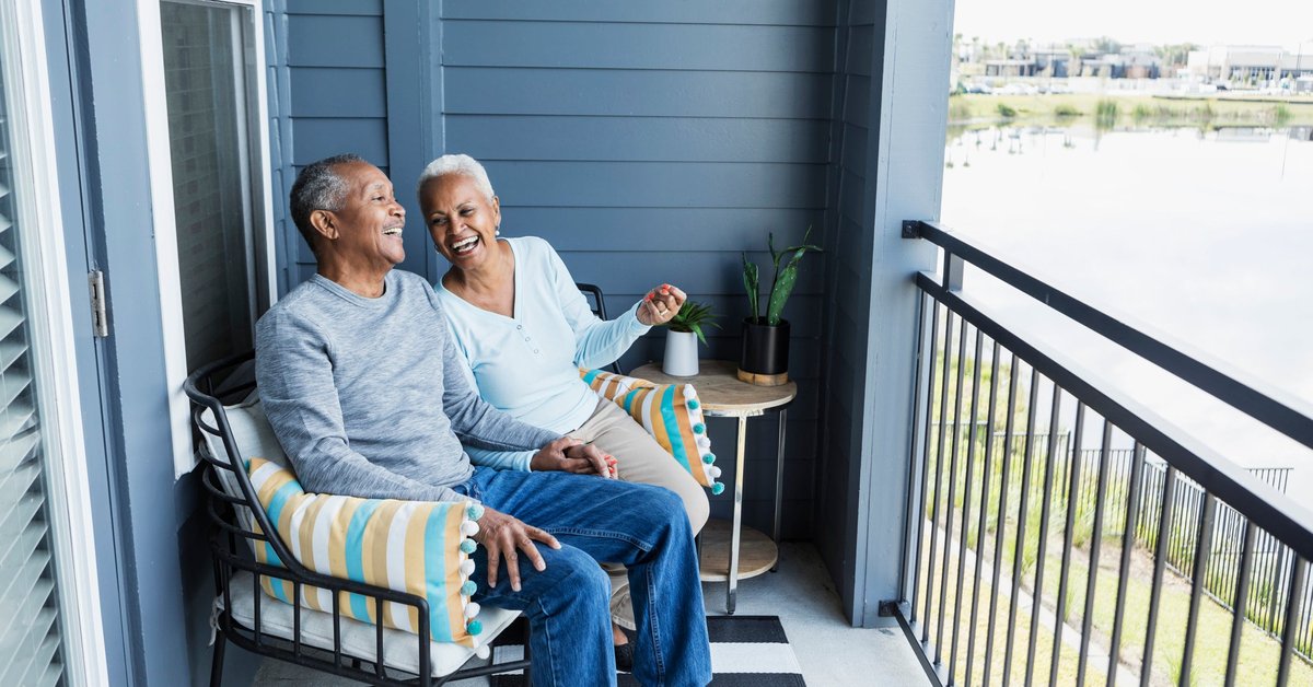 Two mature adults laughing together while sitting on the back deck of their home overlooking a small lake.