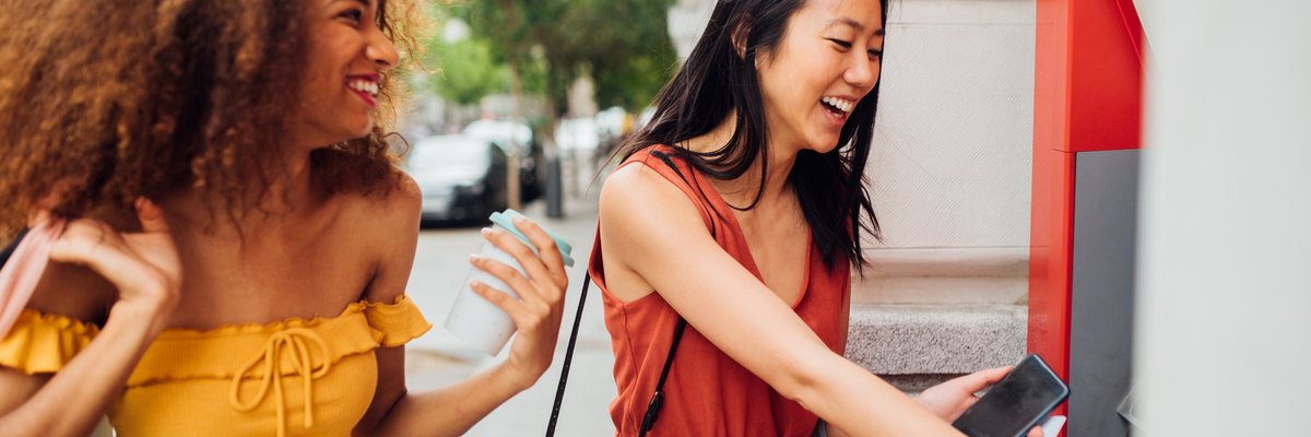 A young woman using an ATM on the street while her friend waits for her.