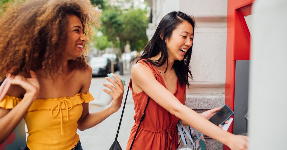 A young woman using an ATM on the street while her friend waits for her.