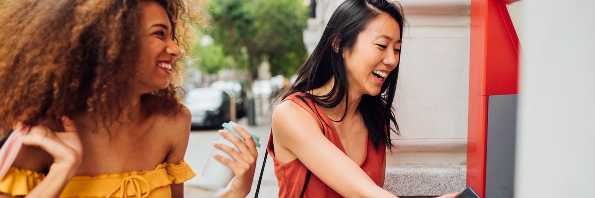 A young woman using an ATM on the street while her friend waits for her.