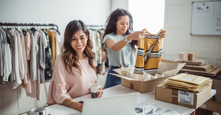 One woman packing clothes into shipping boxes while the other checks her laptop at a table in a clothing store storeroom.