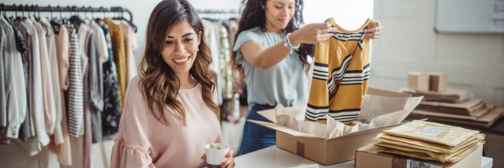 One woman packing clothes into shipping boxes while the other checks her laptop at a table in a clothing store storeroom.