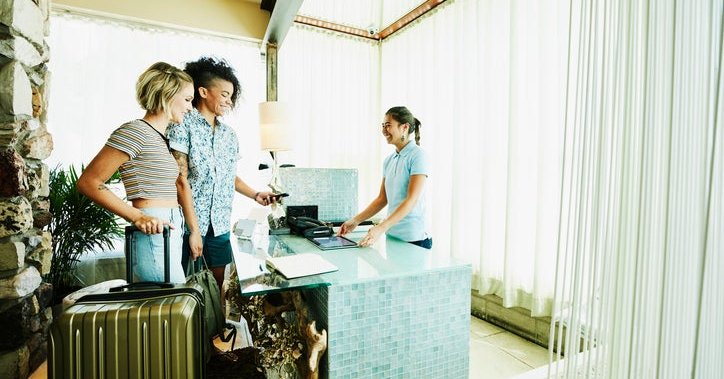 Two people with suitcases checking into a hotel by swiping their phone on the payment reader with an employee behind the lobby desk.