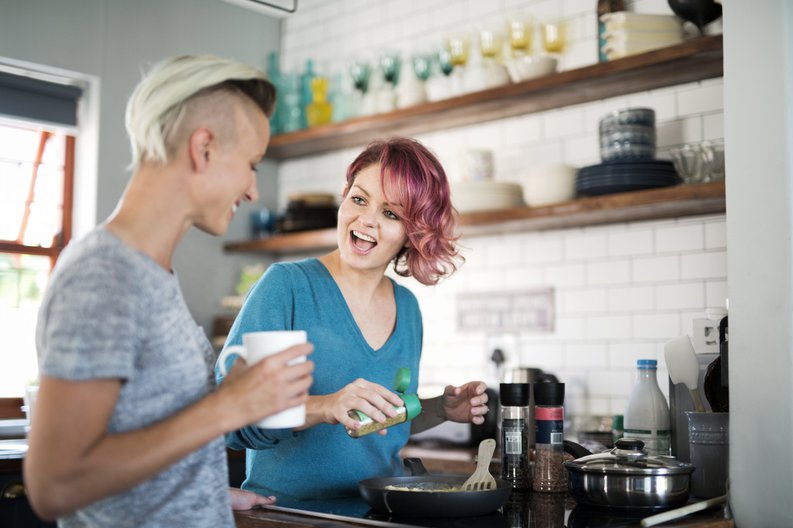 A woman cooking at the stove and laughing with her girlfriend drinking coffee.