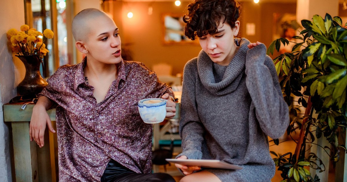 Two women sitting in a cafe while one looks at a tablet in her lap.