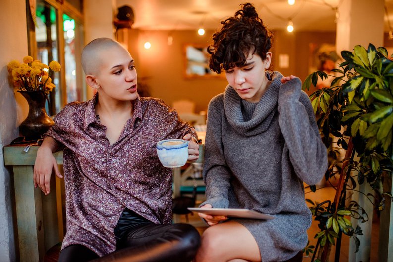 Two women sitting in a cafe while one looks at a tablet in her lap.