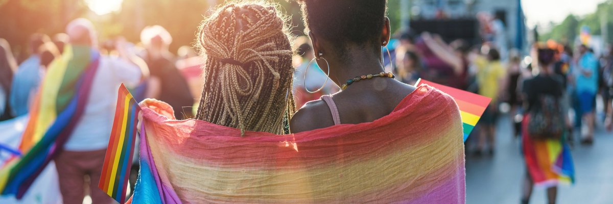 Two women wearing a rainbow flag around their shoulders at a Pride parade.