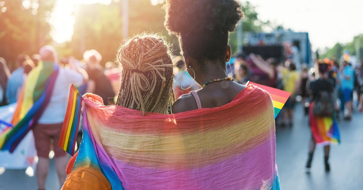 Two women wearing a rainbow flag around their shoulders at a Pride parade.
