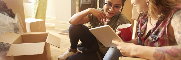 Two women sitting on the floor between moving boxes and looking at something on a tablet one of them is holding.