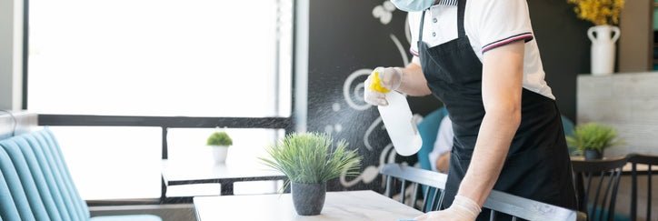 A waiter wearing a medical mask and cleaning a table in a restaurant with a spray bottle and cloth.