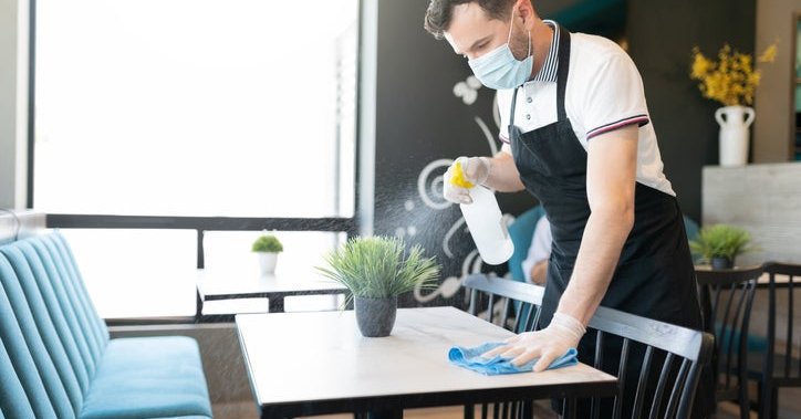 A waiter wearing a medical mask and cleaning a table in a restaurant with a spray bottle and cloth.