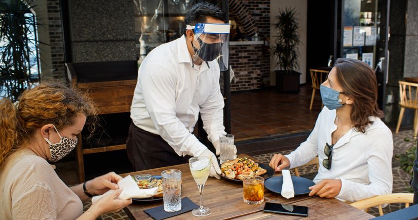 A waiter serving food to two women, all wearing medical masks, at an outdoor dining table.
