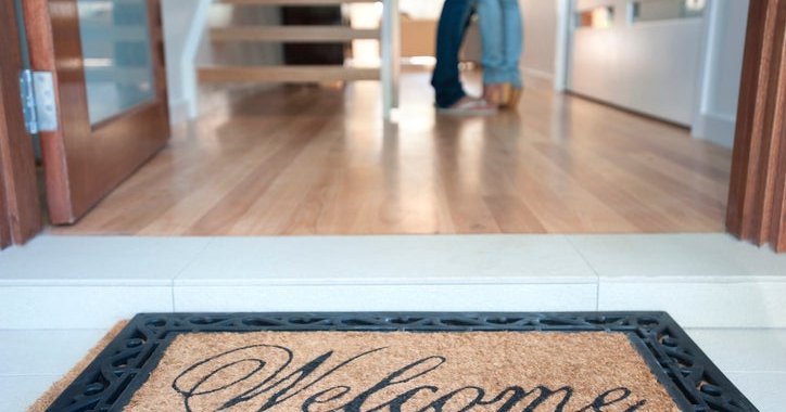 A welcome mat at the open front door of a new house with a man and woman standing inside.