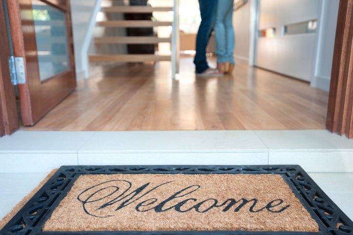 A welcome mat at the open front door of a new house with a man and woman standing inside.