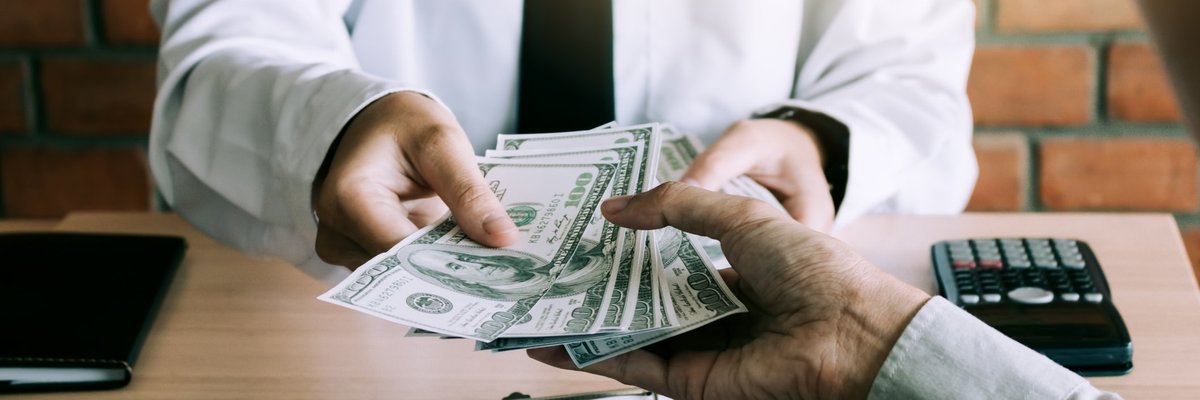 Man in suit handing a stack of hundred dollar bills to a person at a desk.