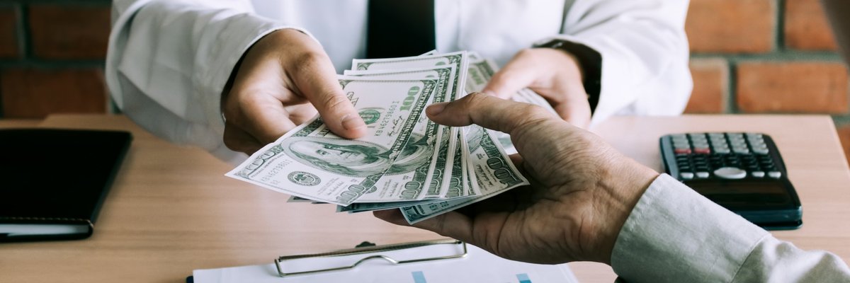 Man in suit handing a stack of hundred dollar bills to a person at a desk.