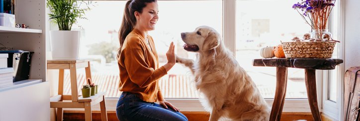 A smiling woman sitting on the hardwood floor of her living room and playing with her dog.