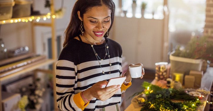 A smiling woman standing at the counter of her store while reading something on her phone.