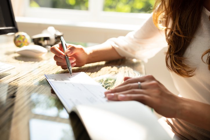 A woman sitting at a sunny desk and signing a check.