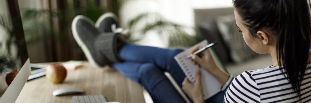A young woman with her slippered feet kicked up on her desk at home and writing in a notebook.