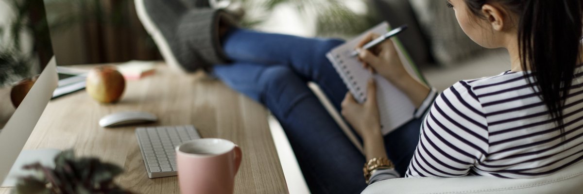 A woman sitting with her feet up on a desk in her home office and writing a to-do list in a notebook.