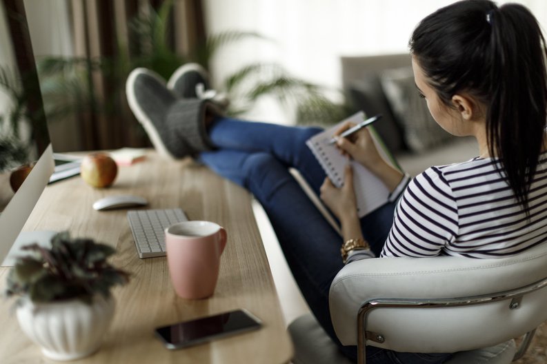 A woman sitting with her feet up on a desk in her home office and writing a to-do list in a notebook.