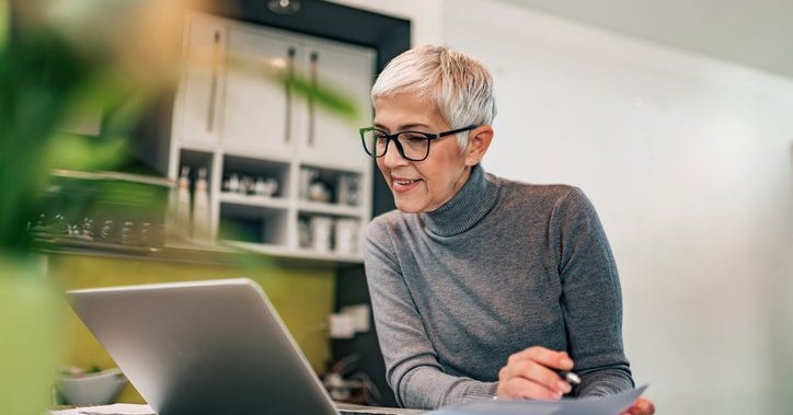 A woman sitting at her kitchen table and looking at a laptop while filling out paperwork.