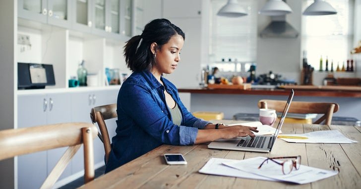 A woman sitting at her kitchen table and typing on a laptop next to paperwork.