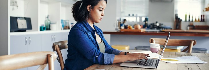 A woman sitting at her kitchen table and typing on a laptop next to paperwork.