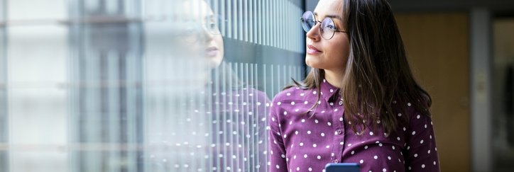 A young professional woman looking thoughtfully out an office window while holding a phone.