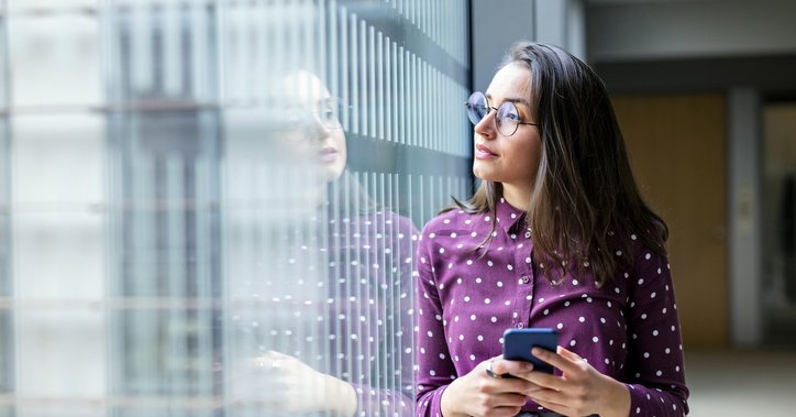 A young professional woman looking thoughtfully out an office window while holding a phone.