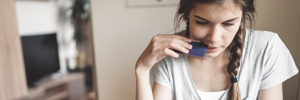 A young woman nervously holding a credit card up to her mouth while looking at her phone.