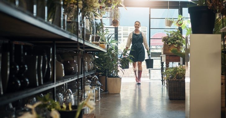 A woman wearing an apron and carrying watering cans into her small business filled with shelves of plants.