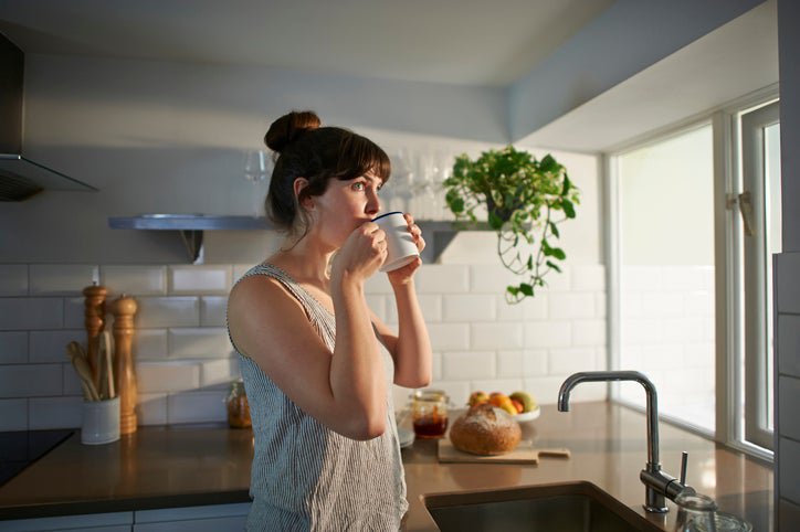 A woman standing next to her kitchen sink while sipping out of a coffee mug.