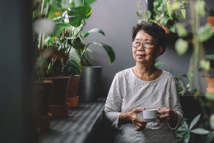 An older woman standing in a room filled with houseplants and drinking tea.