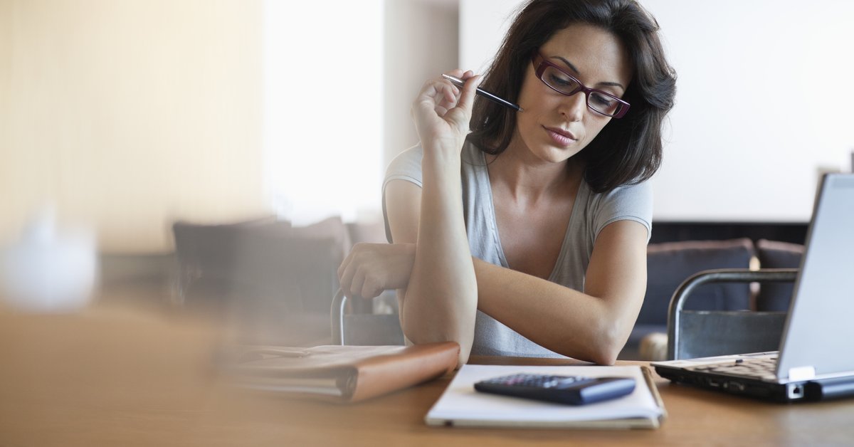 woman sitting at table holding a pen and looking at a calculator and a pad of paper.