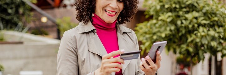 A smiling woman standing outside a sidewalk cafe looking at her phone while holding a credit card.