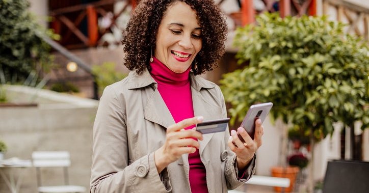 A smiling woman standing outside a sidewalk cafe looking at her phone while holding a credit card.
