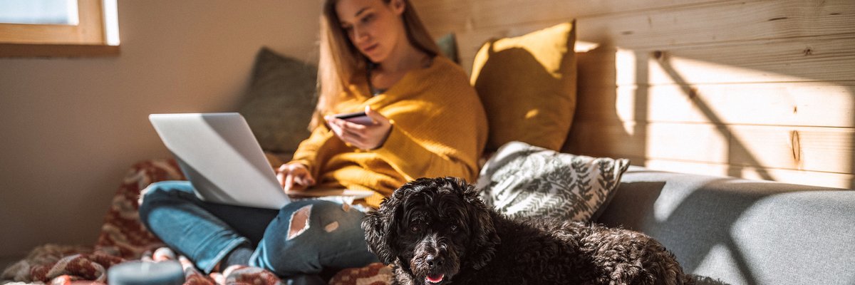 A woman sitting next to her dog on her couch in the sun while holding a credit card and looking something up on her laptop.