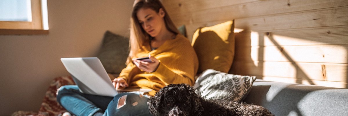 A woman sitting next to her dog on her couch in the sun while holding a credit card and looking something up on her laptop.