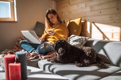 A woman sitting next to her dog on her couch in the sun while holding a credit card and looking something up on her laptop.