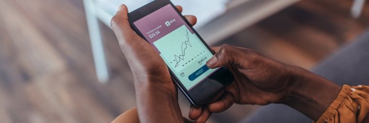A woman sitting on her couch and looking at her phone showing a stock chart on the screen.