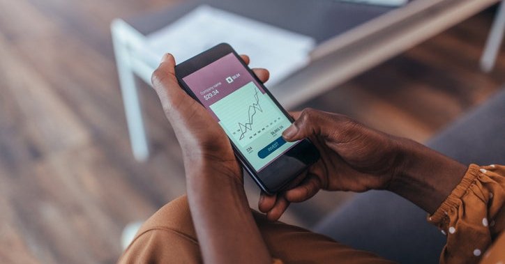 A woman sitting on her couch and looking at her phone showing a stock chart on the screen.
