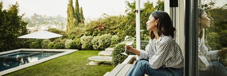 A smiling woman holding a glass of wine while seated on the steps of a patio deck looking at her backyard.