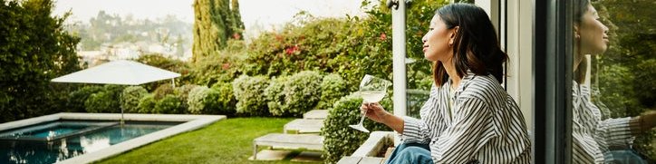 A smiling woman holding a glass of wine while seated on the steps of a patio deck looking at her backyard.