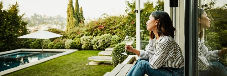 A smiling woman holding a glass of wine while seated on the steps of a patio deck looking at her backyard.