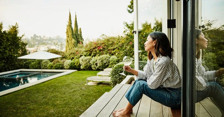 A smiling woman holding a glass of wine while seated on the steps of a patio deck looking at her backyard.
