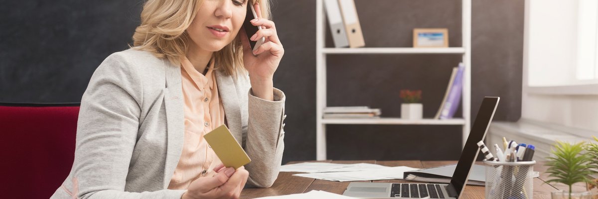 Woman in business suit sitting in front of laptop looking at a credit card and talking on the phone.